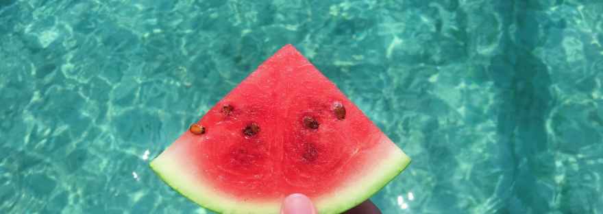 hand holding a slice of watermelon with blue swimming pool water in the background